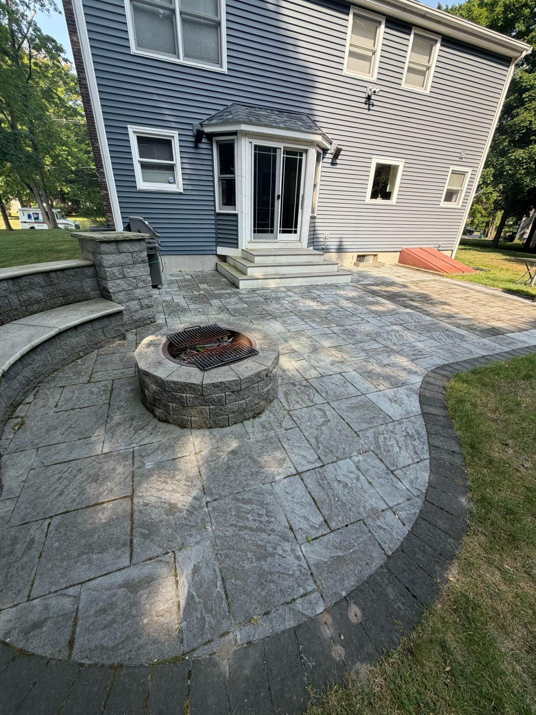 Gray paver patio and fire pit dull with dirt behind a blue house